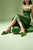 A model in a green satin strapless dress poses with a matching green beaded clutch bag and green embellished slingback heels against a solid sage green studio background, Obroi