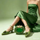 A model in a green satin strapless dress poses with a matching green beaded clutch bag and green embellished slingback heels against a solid sage green studio background, Obroi