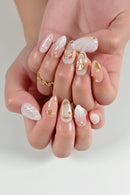 A close-up of hands showcasing a set of long, almond-shaped press-on nails. The nails feature a glossy white base with delicate gold metallic accents, gold leaf details, and small pearl embellishments, Obroi