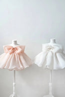 Two sleeveless baby dresses displayed on mannequin stands, one in soft blush pink and one in white, both featuring a large bow at the neckline and a ruffled tutu skirt, Obroi