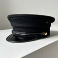 A black military-style peaked cap with a glossy black visor and a small gold button detail, resting on a white surface against a light gray background, Obroi