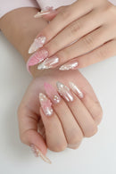 A close-up of manicured fingernails on two hands, featuring glossy almond-shaped nails in pale pink and translucent white with floral, pearl, and metallic embellishments, Obroi