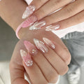 A close-up of manicured fingernails on two hands, featuring glossy almond-shaped nails in pale pink and translucent white with floral, pearl, and metallic embellishments, Obroi