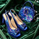 A pair of royal blue slingback peep-toe heels with geometric iridescent patterning and jeweled square embellishments, displayed with a matching blue handbag inside a green velvet box, Obroi