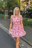 Woman in a pink floral dress standing on a street with greenery and a building in the background, Obroi