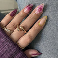 Close-up of a hand with decorative nails and gold rings on a textured surface, Obroi