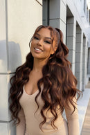 A woman wearing a long, wavy brown hair extension or wig, posing outdoors against a gray stone wall. The brand name 'Obroi' is visible in the top left corner.