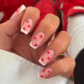 Hand with pink nail polish and red heart designs, wearing a gold ring, with blurred red shoes in the background, Obroi