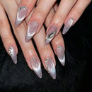 A close-up of manicured hands with long stiletto-shaped nails featuring a silver-to-pink cat-eye gradient and decorative rhinestone and metallic embellishments, set against a black background, Obroi
