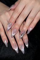 A close-up of manicured hands with long stiletto-shaped nails featuring a silver-to-pink cat-eye gradient and decorative rhinestone and metallic embellishments, set against a black background, Obroi