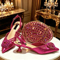 A matching pink high-heeled shoe and embellished oval clutch set displayed on a white round table in an ornate gold interior. The shoes have metallic pink satin uppers with floral crystal embellishments, slingback straps, and kitten heels, Obroi