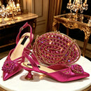 A matching pink high-heeled shoe and embellished oval clutch set displayed on a white round table in an ornate gold interior. The shoes have metallic pink satin uppers with floral crystal embellishments, slingback straps, and kitten heels, Obroi