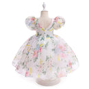 A toddler girl's floral print dress displayed on a dress form mannequin. The dress features a white base with a watercolor-style print of pink, yellow, and blue flowers, green leaves, and crescent moons, complete with puffy short sleeves and a V-neckline, Obroi