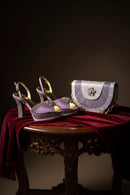 A pair of lavender and gold embellished high-heeled sandals displayed on a dark wooden table draped with deep red velvet, alongside a matching lavender and gold handbag with a jeweled floral clasp, Obroi
