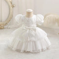 A white toddler girl's party dress displayed on a mannequin dress form. The dress features puffy short sleeves, a lace-trimmed bodice, a large central bow, and a tiered tulle skirt, Obroi