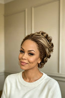 A woman with a styled updo hairstyle featuring highlighted brown and blonde curls, wearing a white knit sweater and gold hoop earrings against a neutral paneled wall, Obroi