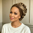 A woman with a styled updo hairstyle featuring highlighted brown and blonde curls, wearing a white knit sweater and gold hoop earrings against a neutral paneled wall, Obroi