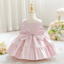 A light pink toddler girl's dress displayed on a mannequin dress form, featuring a large matching bow at the back and a tiered ruffled skirt, set against a soft, neutral indoor background with curtains and flowers, Obroi