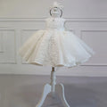 A white toddler girl's formal dress with a matching tiara, displayed on a white dress form mannequin against a white paneled wall background, Obroi