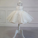 A white toddler girl's formal dress with a matching tiara, displayed on a white dress form mannequin against a white paneled wall background, Obroi