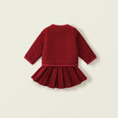 A baby girl's long-sleeve dress in deep red, featuring a textured waffle-knit top and a pleated skirt, shown from the back on a light beige background, Obroi