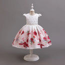 A young girl's white dress displayed on a white mannequin against a solid gray studio background. The dress features a white bodice with tiny silver rhinestone embellishments, a beaded waistband, and a full, flared skirt with a red and pink floral pattern, Obroi