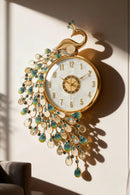 An ornate wall clock with a gold-tone peacock design, turquoise and clear teardrop embellishments, and a white clock face, mounted on a beige wall with strong sunlight casting a shadow, Obroi