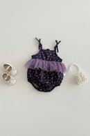 A baby girl's romper in a dark purple floral print with a layered lavender tulle ruffle at the waist, styled in a flat lay with cream baby shoes and a pearl heart bag on a textured off-white background, Obroi