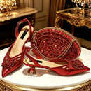 A red satin pointed-toe slingback high heel with a jeweled floral embellishment, shown beside a matching red embellished clutch and a second shoe on a white marble table with ornate gold surroundings, Obroi