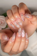 A close-up of hands showcasing almond-shaped press-on nails with a glossy pink and nude gradient, decorated with white butterfly and floral designs, delicate rhinestones, and a subtle glitter finish, Obroi