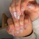 A close-up of hands showcasing almond-shaped press-on nails with a glossy pink and white gradient, decorated with white butterfly and floral designs, delicate rhinestone accents, and a subtle glitter finish, Obroi