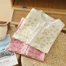 Two folded fabric items, one white with gold patterns and one pink, on a woven surface, Obroi