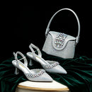 A coordinated evening accessories set featuring a silver rhinestone-embellished handbag and matching pointed-toe slingback heels, displayed on a dark green velvet surface against a black background, Obroi