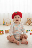 Child wearing a red beret with strawberry designs, sitting in a room with a teddy bear in the background, Obroi