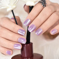 Hand with purple glittery nails holding a nail polish bottle with white flowers in the background, Obroi