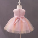 A sleeveless pink children's party dress displayed on a white dress form mannequin against a solid dark gray background. The dress features a structured pink bodice, a large matching pink bow at the back waist, and a layered tulle skirt with a soft peach and pale yellow gradient, Obroi