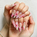 A close-up of a hand displaying long, sculpted stiletto nails with intricate pink and white floral, polka dot, and wave designs, adorned with small gold rhinestones, Obroi