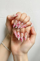 A close-up of a hand displaying long, sculpted stiletto nails with intricate pink and white floral, polka dot, and wave designs, adorned with small gold rhinestones, Obroi