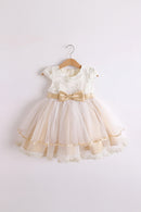 A toddler girl's formal party dress hanging on a wooden hanger against a plain light background. The dress features a cream lace bodice with gold floral embroidery, short sleeves, a beige satin waistband with a large bow, and a layered tulle skirt in cream and beige with gold ribbon trim, Obroi