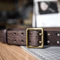 A close-up of a dark brown textured leather belt with a square antique brass buckle, resting on a wooden surface next to a cutting mat, Obroi
