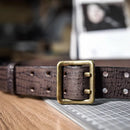 A close-up of a dark brown textured leather belt with a square antique brass buckle, resting on a wooden surface next to a cutting mat, Obroi