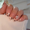 A close-up of a hand with long almond-shaped nails decorated with white and pink floral designs, black leaf accents, and thin gold lines, resting on a white surface, Obroi