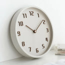 A round wall clock with a textured off-white face and rim, featuring dark brown wooden numerals and hands, shown leaning against a white wall on a white surface with a small stack of books, Obroi