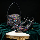 A luxury handbag and matching pointed-toe slingback heels displayed together on a draped green velvet surface with a gold-trimmed white pedestal against a black background. The items are heavily embellished with multicolored rhinestones and black leather, with gold-tone hardware, Obroi