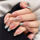 A close-up of a hand with long almond-shaped nails featuring a glossy nude-beige gradient, light blue French tips, and one nail with a subtle geometric pattern, Obroi
