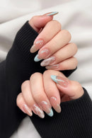 A close-up of a hand with long almond-shaped nails featuring a glossy nude-beige gradient, light blue French tips, and one nail with a subtle geometric pattern, Obroi