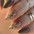 A close-up of a hand with manicured nails, featuring two almond-shaped nails with a glittery taupe leopard print pattern and two glossy taupe nails, accessorized with gold rings, Obroi