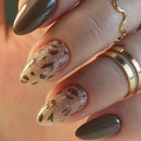 A close-up of a hand with manicured nails, featuring two almond-shaped nails with a glittery taupe leopard print pattern and two glossy taupe nails, accessorized with gold rings, Obroi