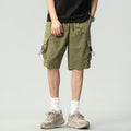 A person wearing olive green cargo shorts, a black t-shirt, white socks, and beige sneakers, shown from the waist down against a light gray studio background, Obroi