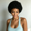A woman with short, curly black hair wearing a light blue ribbed tank top and gold hoop earrings, looking down with a soft smile against a plain light gray background, Obroi